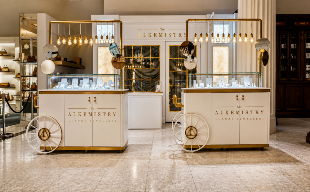 The Alkemistry luxury jewellery pop-up at Selfridges London featuring bespoke white and gold display carts, illuminated pendant lighting, glass showcases and a premium retail environment designed and delivered by NORTHBANKS.